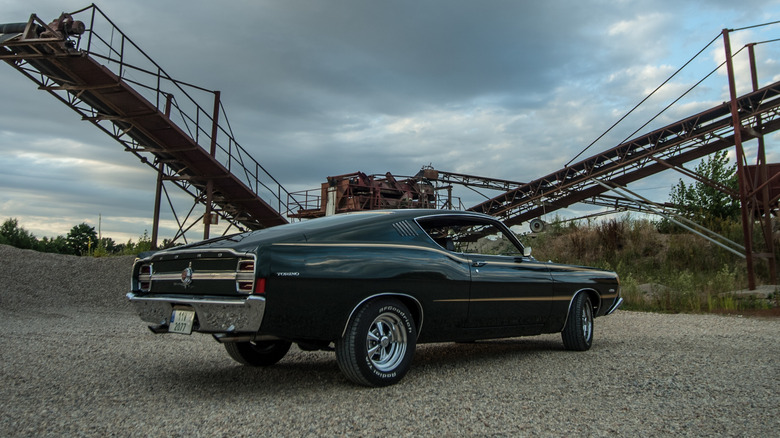 Rear 3/4 view of a dark green Ford Torino against an industrial backdrop