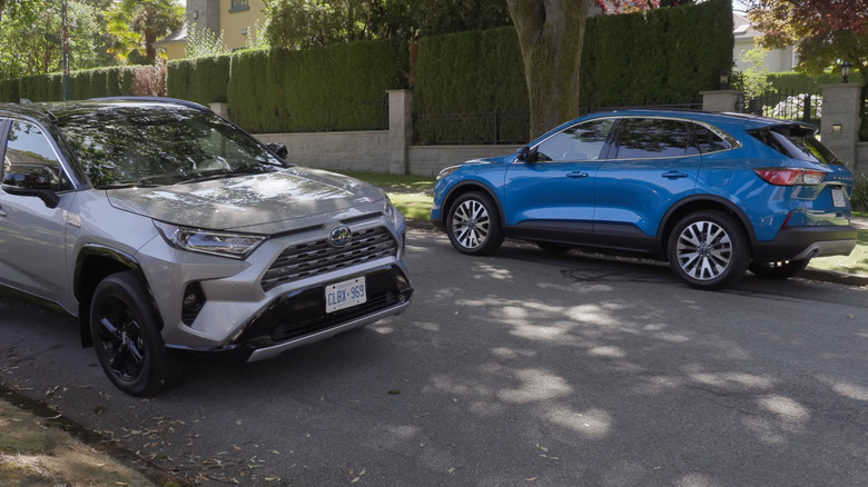 A blue Ford Escape from 2021 and a silver 2021 Toyota RAV4 standing opposite each other on a suburban road in the shade.