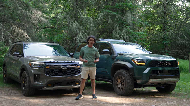 A 2026 Toyota 4Runner finished in dark green standing in a forest clearing next to a beige 2026 Ford Explorer.