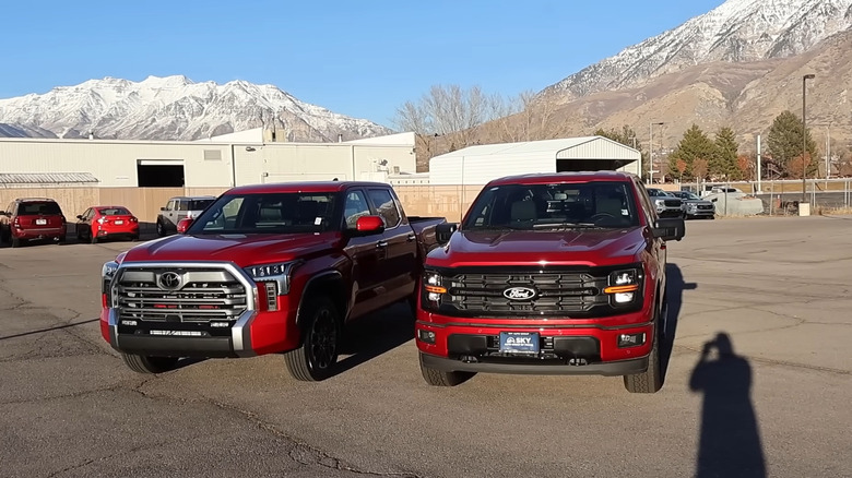 A 2026 Ford F-150 and a 2026 Toyota Tundra truck, both finished in metallic red, standing in a paved lot with a snowy mountain in the back.