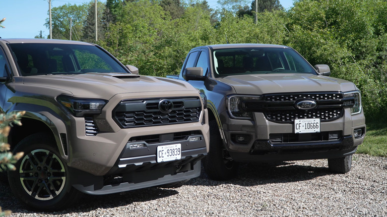 A brown 2026 Ford Ranger and a 2026 Toyota Tacoma standing together on a gravel road, front view visible.