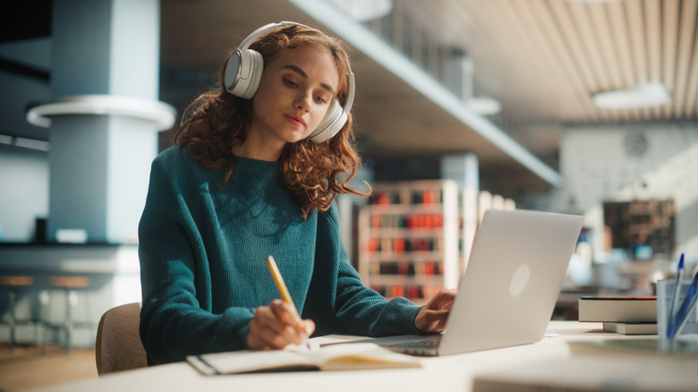 A student with a laptop wearing headphones while writing in a notebook