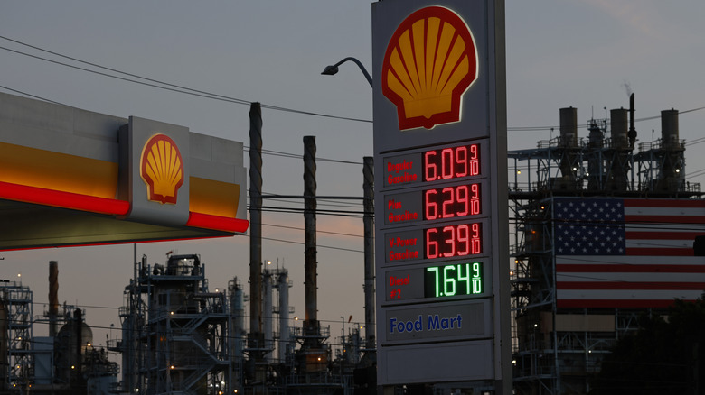 A Shell gas station display showing gas prices, with factories in the background.