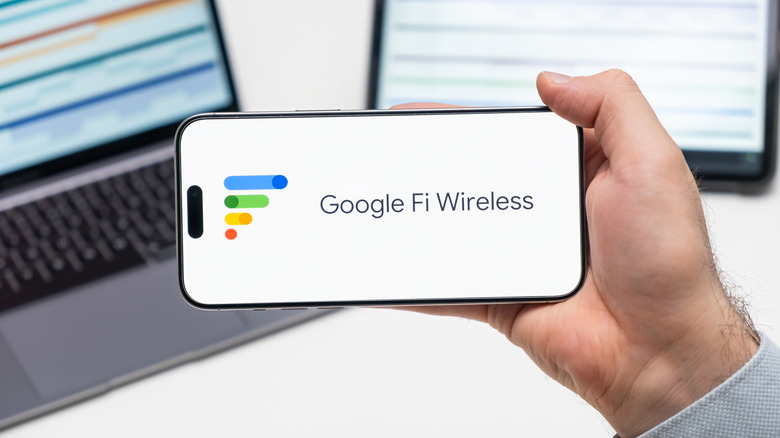 Google Fi Wireless logo of app on the screen of mobile phone held by man in front of the laptop and tablet