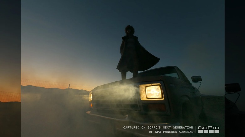 A person standing on the hood of a car with lit headlights at twilight.