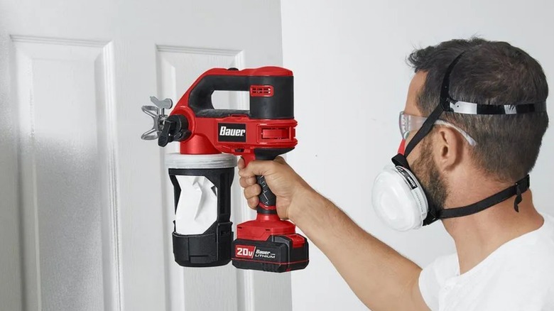 A man wearing mask using red color Bauer paint sprayer on a door
