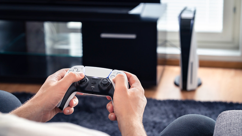 person holding DualSense controller with a PS5 console in the background