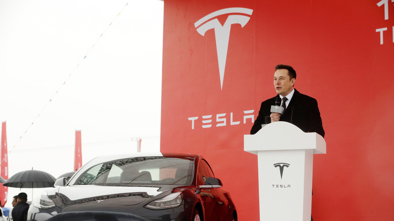 Elon Musk holding a mic in front of a Tesla logo 