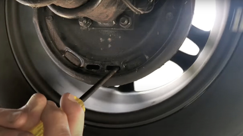 A hand holds a screwdriver inside a slot on the backing plate of a drum brake and wheel.