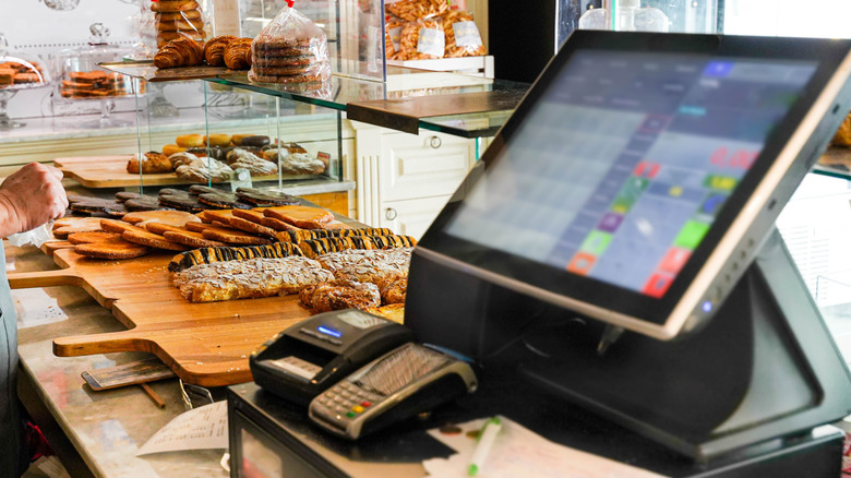 POS (Point of sale) equipment in a bakery. The cash register and other POS equipment is blurred in the foreground while bakery products like croissants and cookies are in focus in the background.