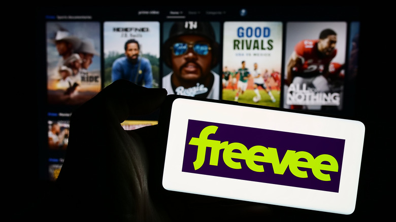 A person holding a smartphone with the Freevee logo on it and a big-screen device in the background with Freevee content.