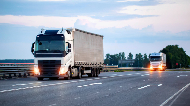 A column of trucks with semi-trailers and headlights on is transporting cargo along the road in the evening
