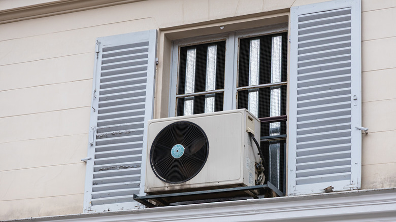 An air conditioner hangs from a window of a building, helping to cool the interior on a sunny summer day. The shutters are partially open.