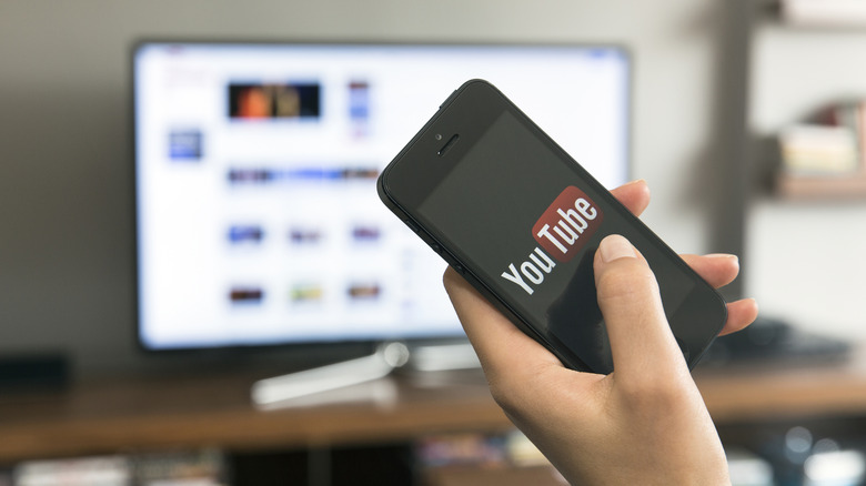 A person holding and touching Apple iPhone 5 that's displaying log in screen of YouTube application with a background of a TV