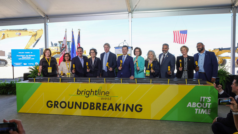 A panel of officials, including former Secretary of Transportation Pete Buttigieg, behind a banner for Brightline West's groundbreaking ceremony in Nevada.