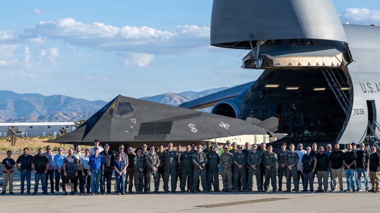 A group of people standing before a C-5M Super Galaxy as an F-117A Nighthawk is loaded inside