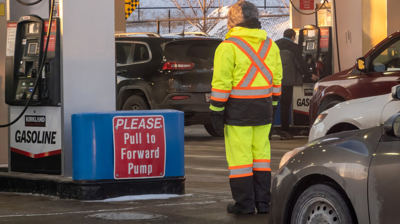 A Costco gas station attendant in high-visibility gear stands near a pump, ensuring vehicles pull forward as others refuel on a cold winter day.