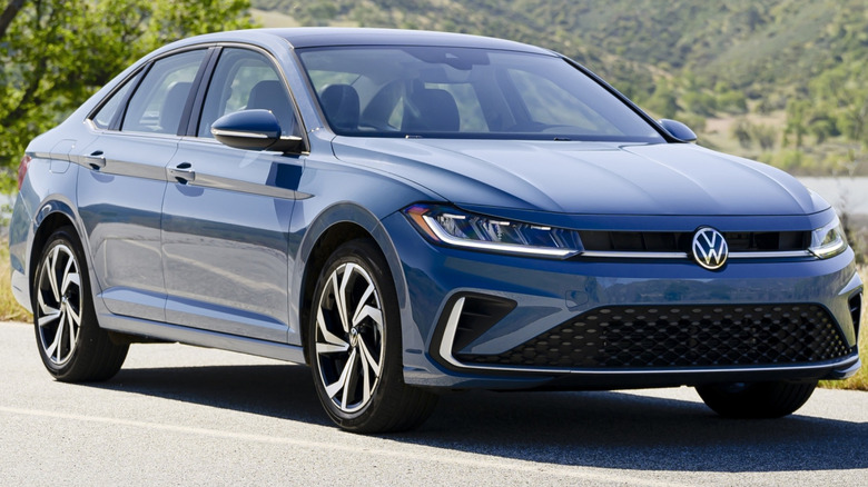A blue 2025 Volkswagen Jetta on a highway with trees and hills in the background.