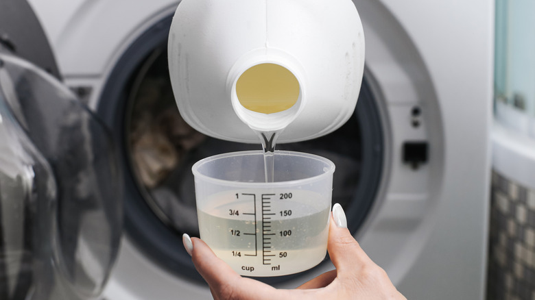 Liquid detergent falls into a cup from a bottle in front of a washing machine