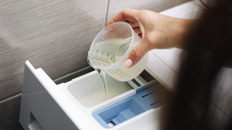 PoA hand pours cup of liquid detergent into a washing machine drawer.