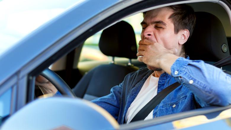 A person yawning, eyes closed, while sitting behind the steering wheel of their car.