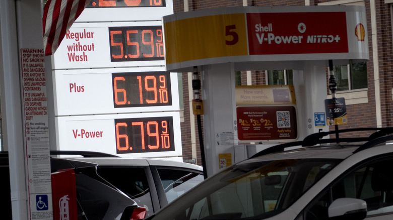 Shell gas station with cars in foreground, focusing on sign reading $5.59 for Regular, $6.19 for Plus, $6.79 for V-Power