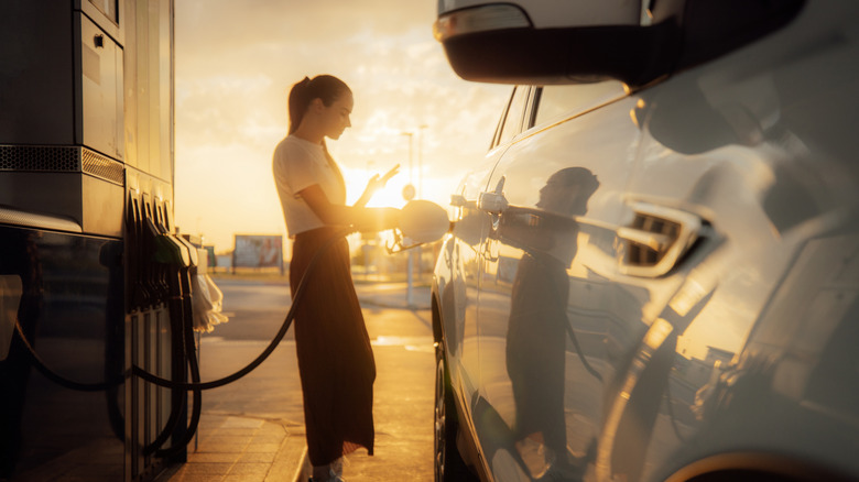 Woman filling up generic SUV at gas pump during sunset, figure silhouetted by sun