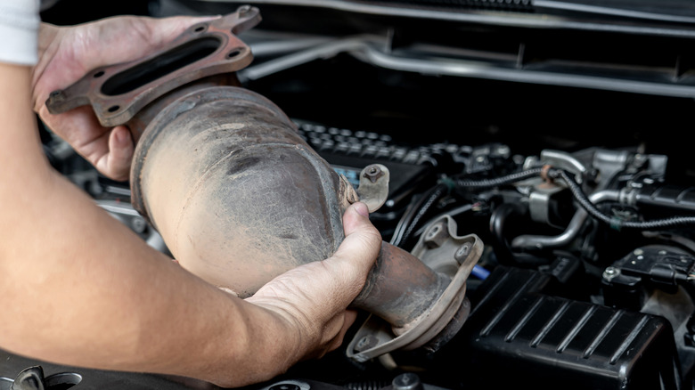 Close-up of an old catalytic converter help in hand by a person above the engine bay.