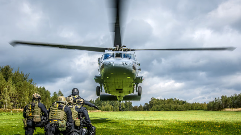 Black Hawk helicopter low over grass field with soldiers waiting in front