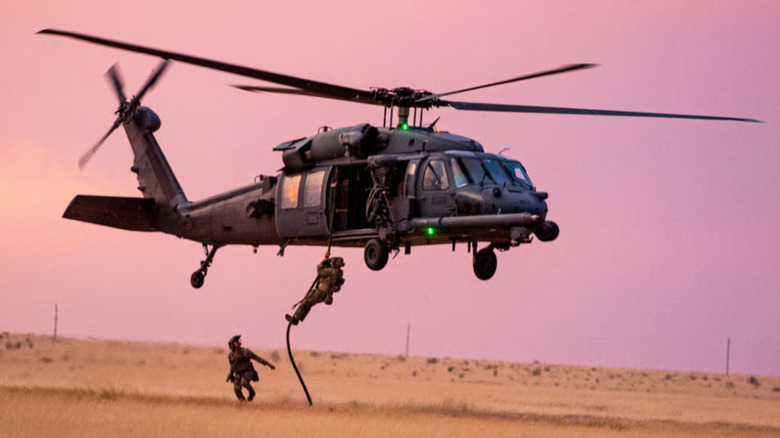 Soldier dropping from rope beneath Black Hawk helicopter in dry field at dusk
