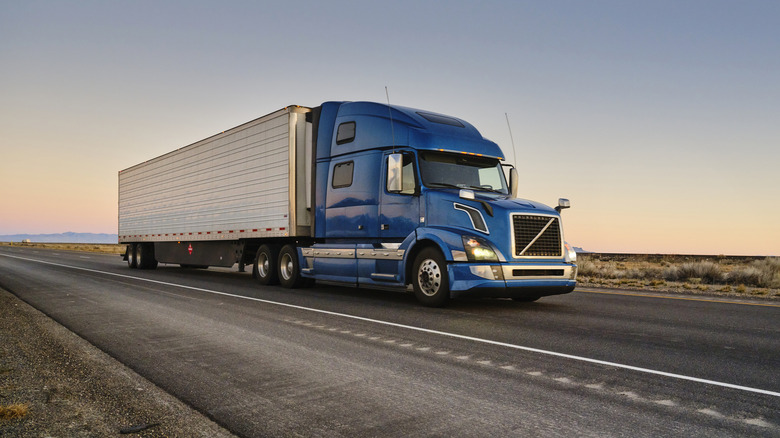 Blue semi-truck driving on a desert road at sunset