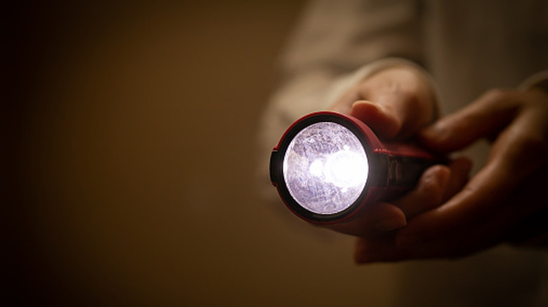 A focus on a red colored flashlight emitting a white beam as a person holds it in their hands