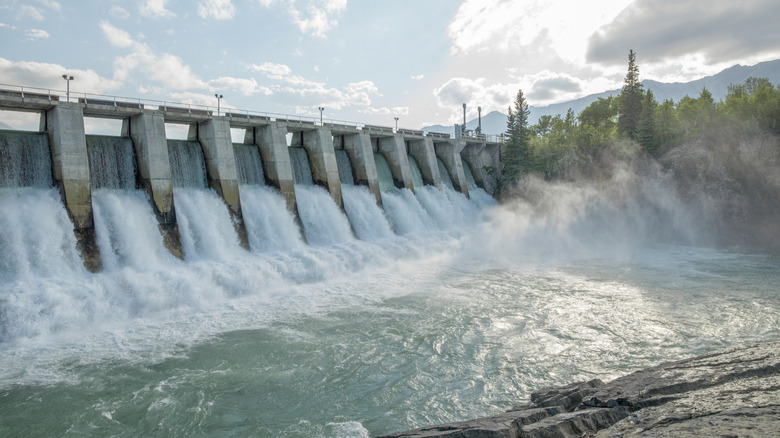 A large dam with water rushing through it, with trees and mountains in the background