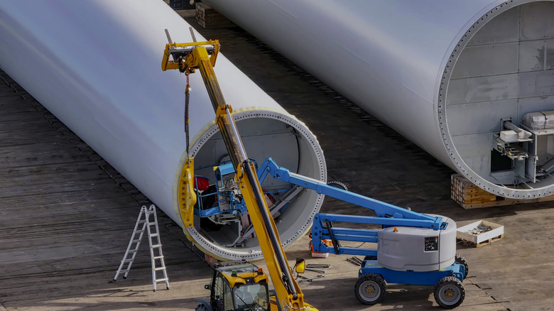 Massive wind turbine towers lying on a dockside with two cranes working on them.