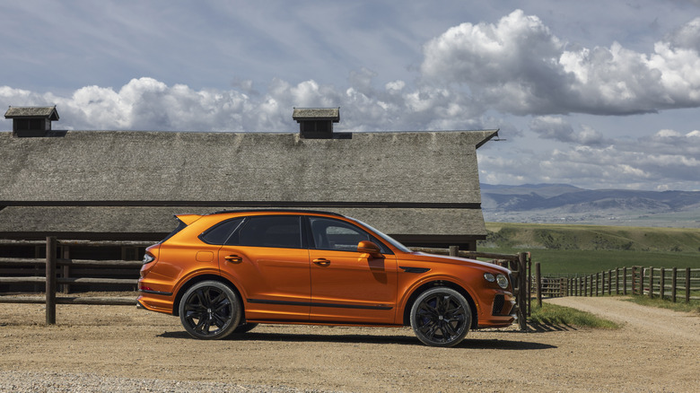 Side shot of an orange Bentley Bentayga next to a famhouse