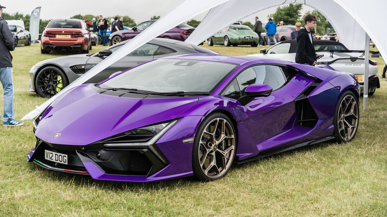A purple Lamborghini Revuelto on display, parked on grass alongside other high-end sports cars, front 3/4 view