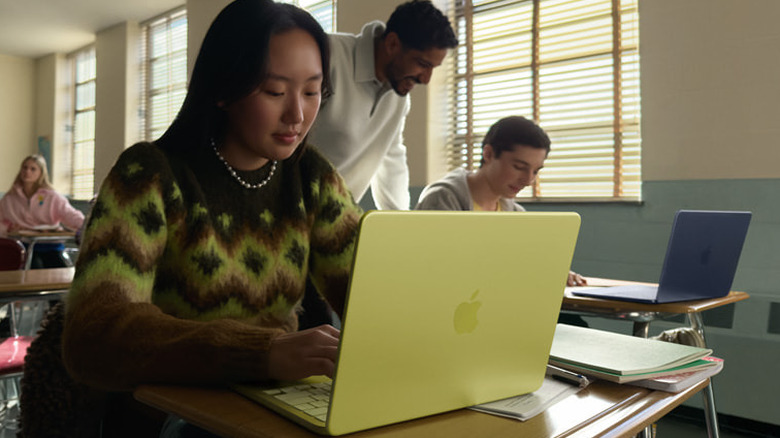 A person using a yellow MacBook Neo on a desk in a classroom, with other people behind her