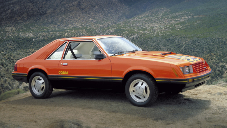 An orange 1979 Ford Mustang Cobra with black trim parked on a cliff with mountains in the background.
