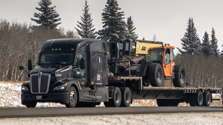 A black Kenworth T680 semi-truck hauling a telehandler on a flatbed trailer along a highway.