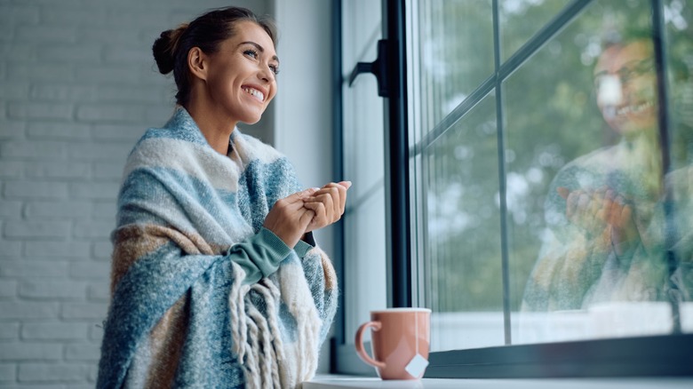 Smiling woman with tea and blanket on shoulders looking out window