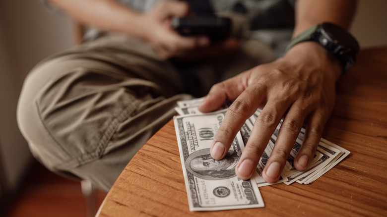 A person holding a few hundred dollars bills against the table while typing on a cell phone