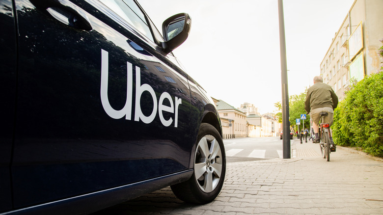 A black vehicle with an Uber sticker parked on a sidewalk with a person riding a bicycle near it.