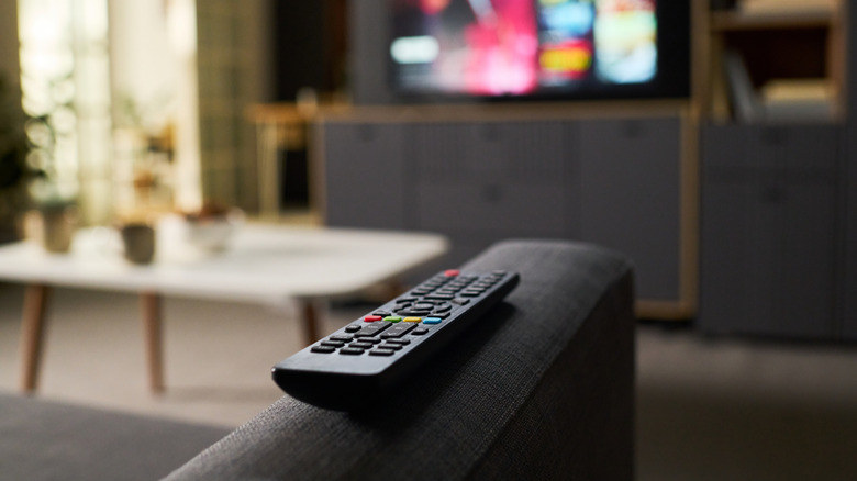 A remote control sitting on a gray couch armrest, TV displaying something in the background