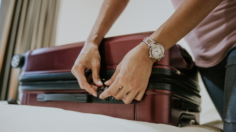 A person wearing a watch locking a maroon suitcase.