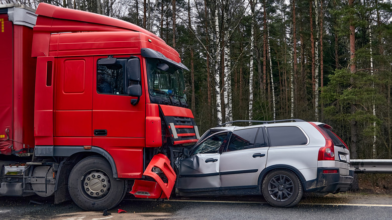 A red semi-truck has collided with a silver SUV on a forest-lined road.