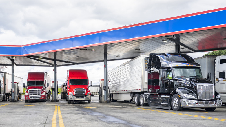 Several semi trucks parked at a truck stop fuel station under a blue and red canopy on a cloudy day.