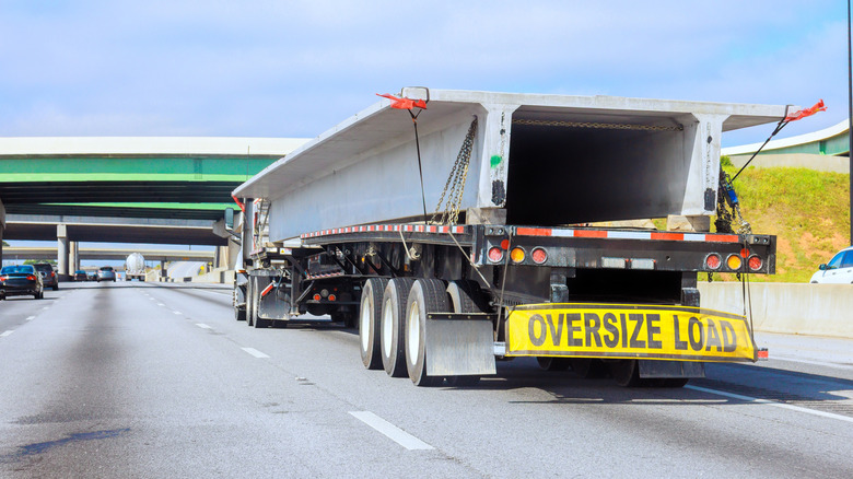 A semi-truck hauling an oversized concrete beam on a flatbed trailer travels down a multi-lane highway, marked with a yellow "OVERSIZE LOAD" banner.