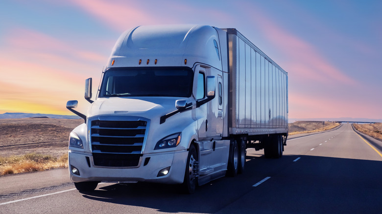 A white semi-truck with a sleeper cab and enclosed trailer driving on an open highway at sunset.