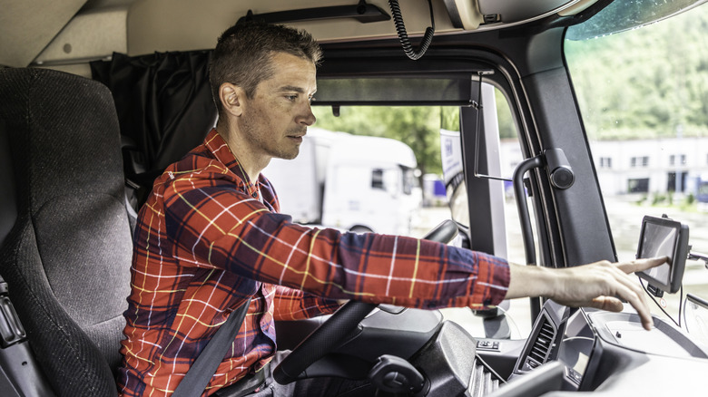 A truck driver wearing a red plaid shirt sits in the cab of a semi-truck, focused as he reaches to operate the GPS navigation system mounted on the dashboard.
