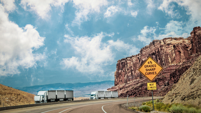 Several semi-trucks sit next to a rocky desert canyon road, with a sign warning of 6% grades and sharp curves for the next 3 miles.